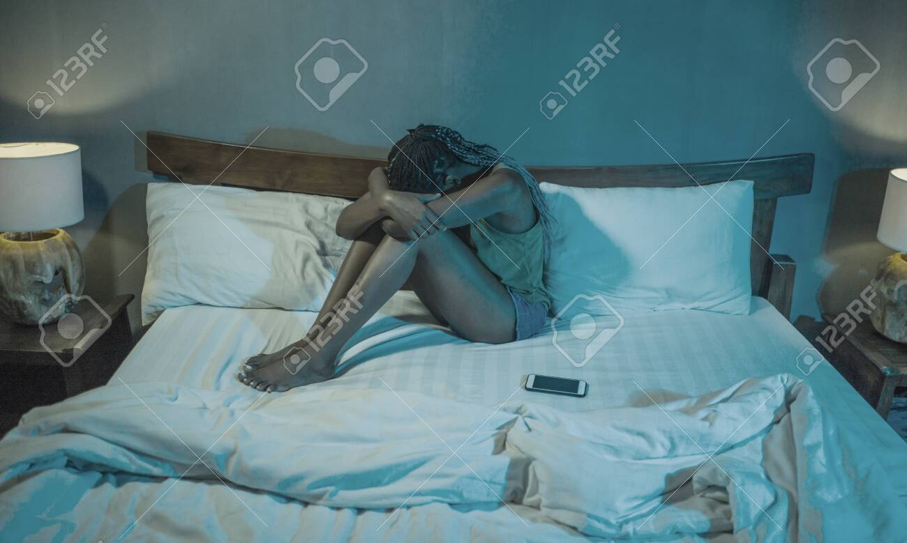 dramatic lifestyle portrait of young sad and depressed black African American woman crying in bed with mobile phone victim of cyber bullying or broken heart in her bedroom - 148152132 dramatic lifestyle portrait of young sad and depressed black African American woman crying in bed with mobile phone victim of cyber bullying or broken heart in her bedroom - 148152132