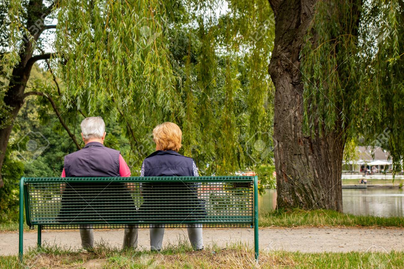 Senior couple on a bench in a park at a lake, shot from behind - 128676526 Senior couple on a bench in a park at a lake, shot from behind - 128676526
