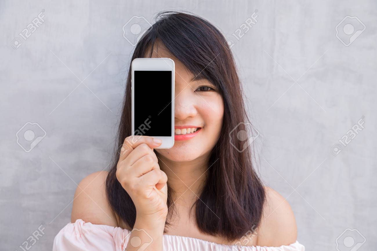 Smiling woman showing a blank smartphone screen standing on concrete wall. - 60266235 Smiling woman showing a blank smartphone screen standing on concrete wall. - 60266235