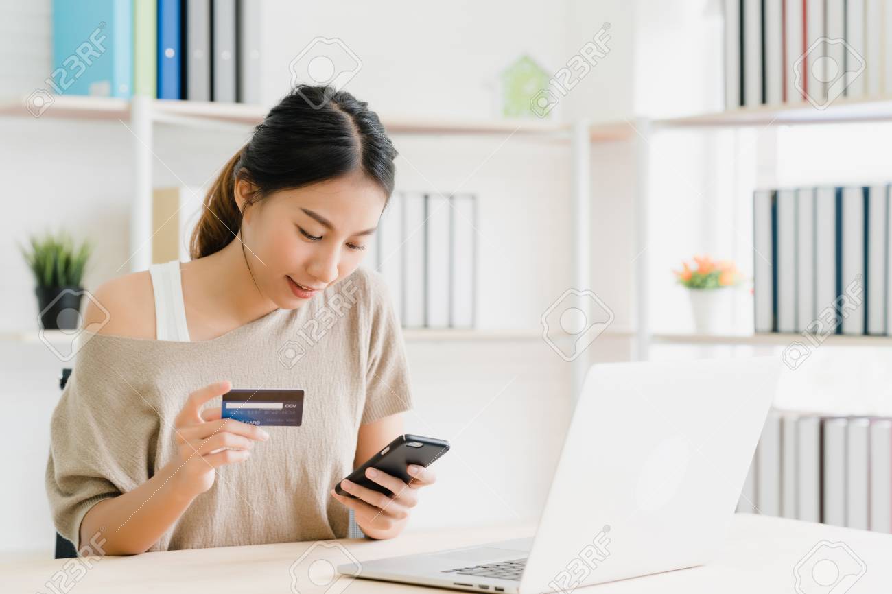 Beautiful Asian woman using smartphone buying online shopping by credit card while wear sweater sitting on desk in living room at home. Lifestyle woman at home concept. - 120849404 Beautiful Asian woman using smartphone buying online shopping by credit card while wear sweater sitting on desk in living room at home. Lifestyle woman at home concept. - 120849404