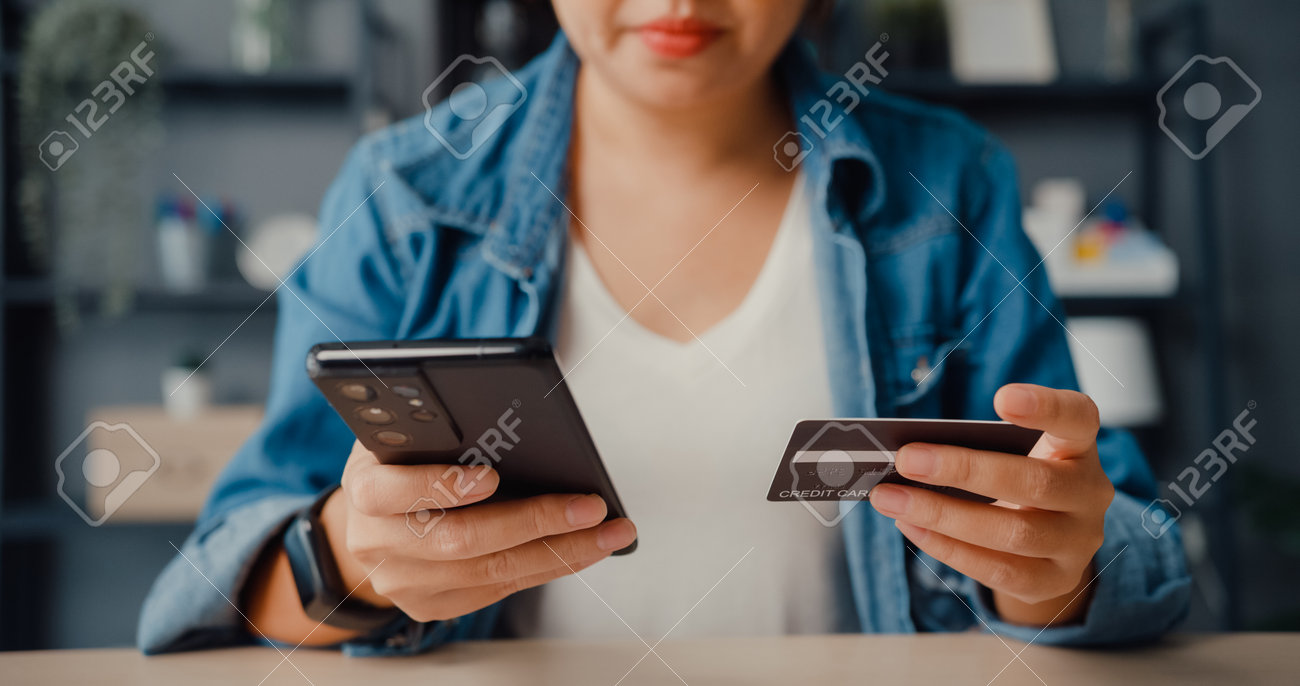 Young asia lady use cellphone order online shopping product and paying bills with credit card in living room interior. Stay at house, Self quarantine activity, Fun activity for coronavirus prevention. - 169421863 Young asia lady use cellphone order online shopping product and paying bills with credit card in living room interior. Stay at house, Self quarantine activity, Fun activity for coronavirus prevention. - 169421863