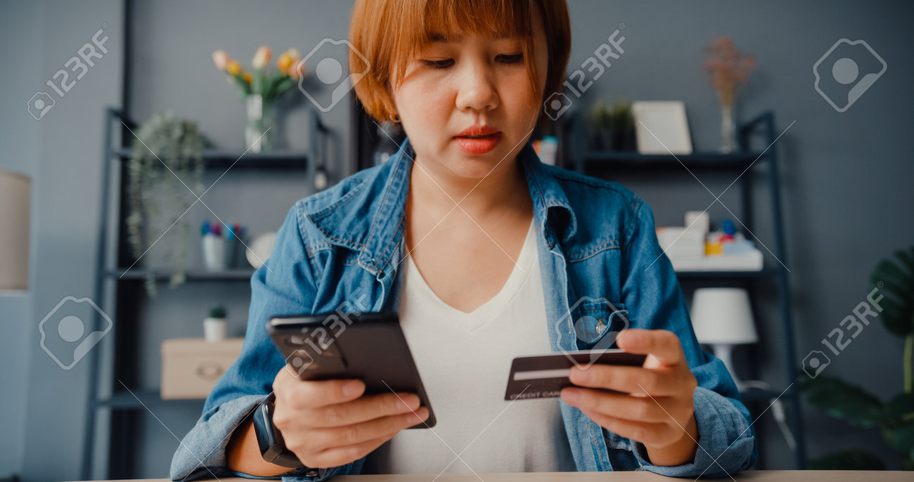 Young asia lady use cellphone order online shopping product and paying bills with credit card in living room interior. Stay at house, Self quarantine activity, Fun activity for coronavirus prevention. - 169421869 Young asia lady use cellphone order online shopping product and paying bills with credit card in living room interior. Stay at house, Self quarantine activity, Fun activity for coronavirus prevention. - 169421869