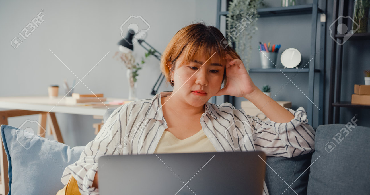 Freelance Asia lady feel headache while sitting on couch with laptop online learn in living room at house. Working from home, remotely work, social distancing, quarantine for corona virus prevention. - 169422738 Freelance Asia lady feel headache while sitting on couch with laptop online learn in living room at house. Working from home, remotely work, social distancing, quarantine for corona virus prevention. - 169422738