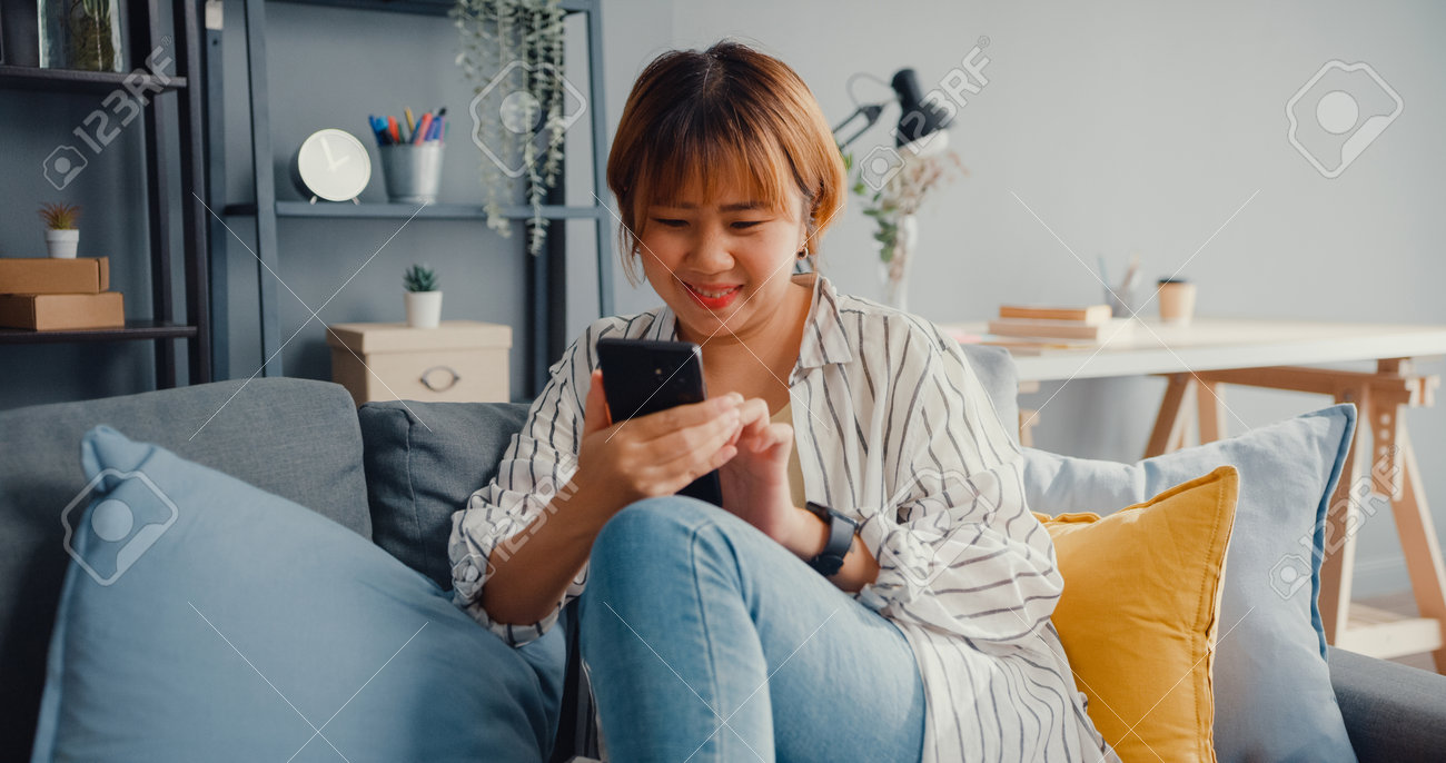 Young Asia lady using smartphone text message or check social media on sofa in living room at house. Work from home, play game, shopping online, social distance, quarantine for coronavirus prevention. - 169422740 Young Asia lady using smartphone text message or check social media on sofa in living room at house. Work from home, play game, shopping online, social distance, quarantine for coronavirus prevention. - 169422740