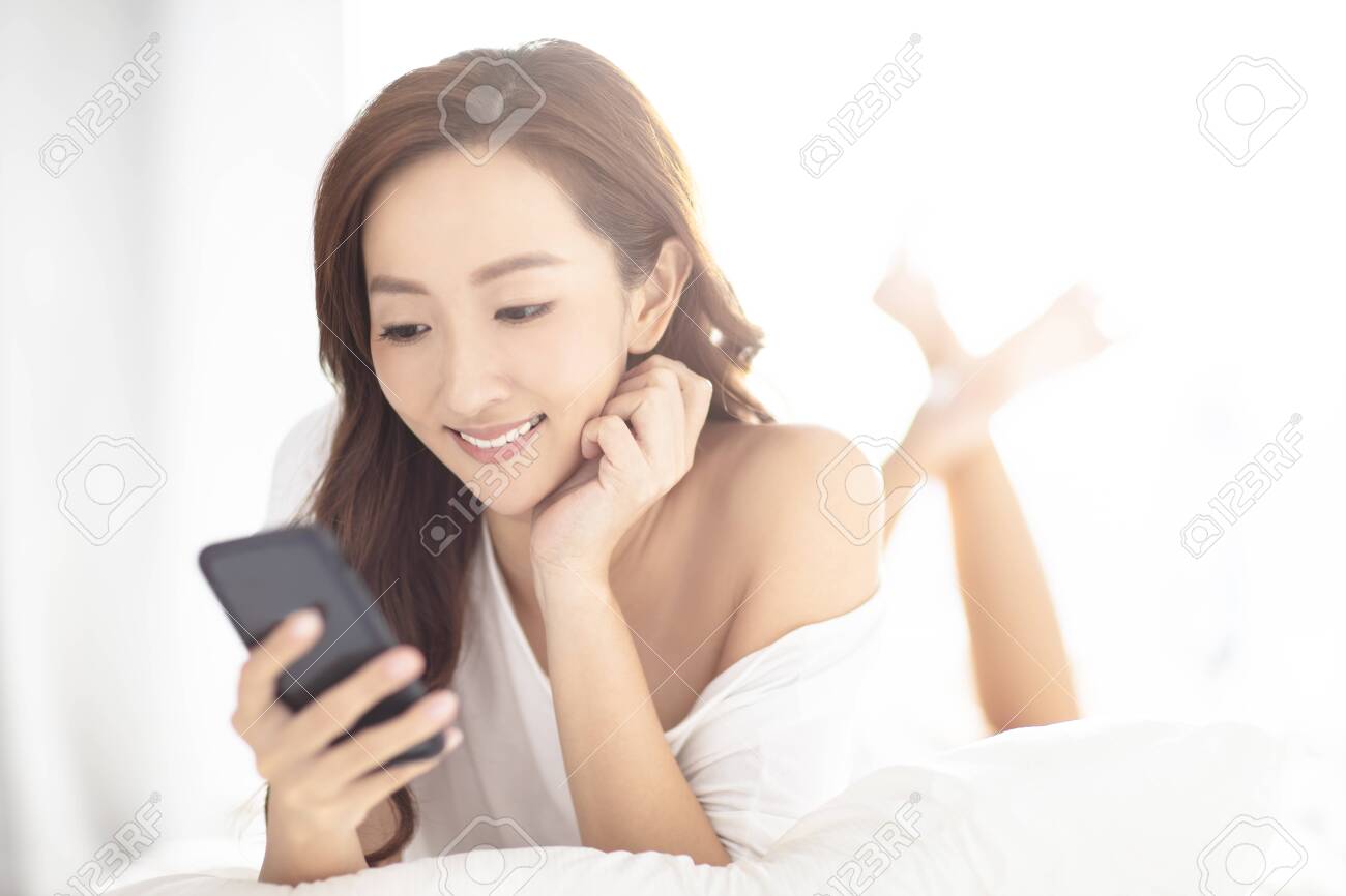 young smiling woman watching mobile phone on bed - 124510003 young smiling woman watching mobile phone on bed - 124510003