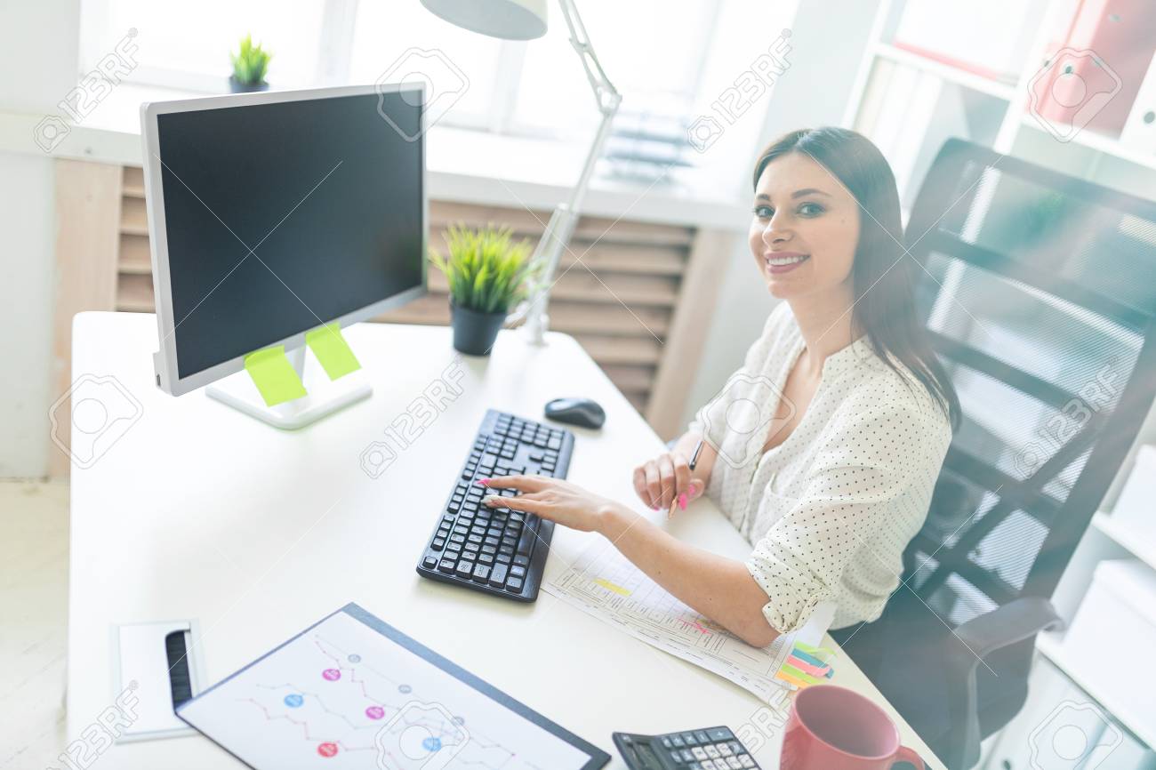 Beautiful Young Girl Working In A Bright Office At A Computer Desk. She Holds In Her