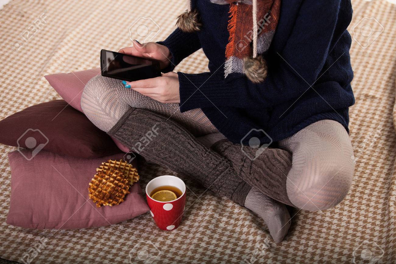 床とタブレットの使用の上に座っている若い女性 居心地の良い家庭的な雰囲気 居心地の良い靴下を編んだ服を着て足 一杯のコーヒーを保持している女性 の写真素材 画像素材 Image