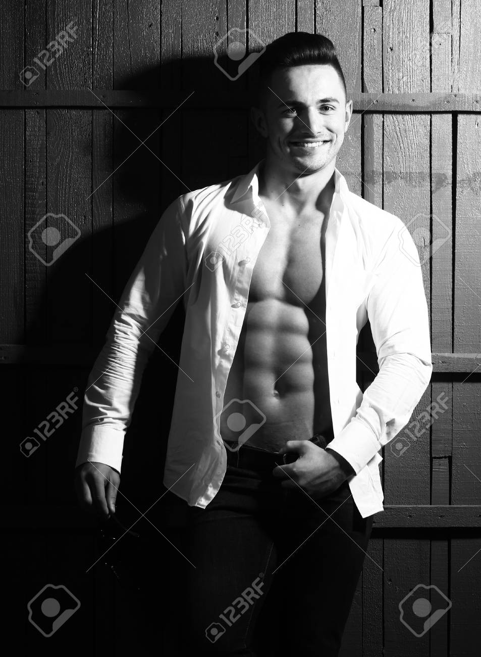 セクシーなおしゃれな屋内木製の背景画像の垂直方向に立っている白いシャツで裸の胴体と官能的な筋肉の若いマッチョな男の笑みを浮かべて の写真素材 画像素材 Image
