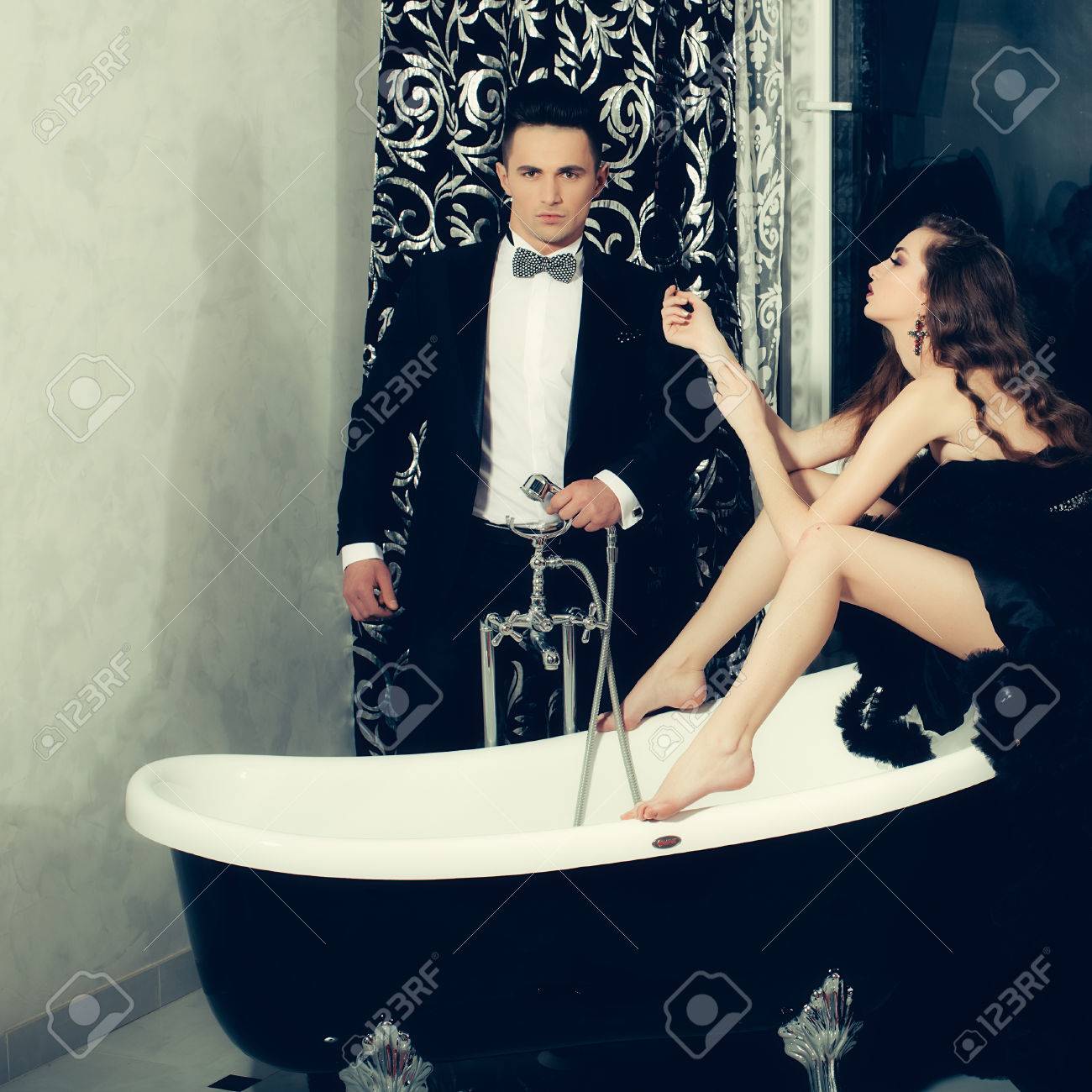 スカートとお風呂に座って長い巻き毛があるエレガントなイブニング ドレスで非常にセクシーな女性と黒のスーツと弓で若いハンサムな男 の写真素材 画像素材 Image