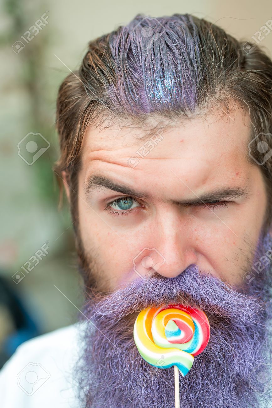 若いハンサムなひげを生やした長いひげ青い色とカラフルなキャンディー クローズ アップを食べる白いシャツで感情的な顔を持つ男 の写真素材 画像素材 Image