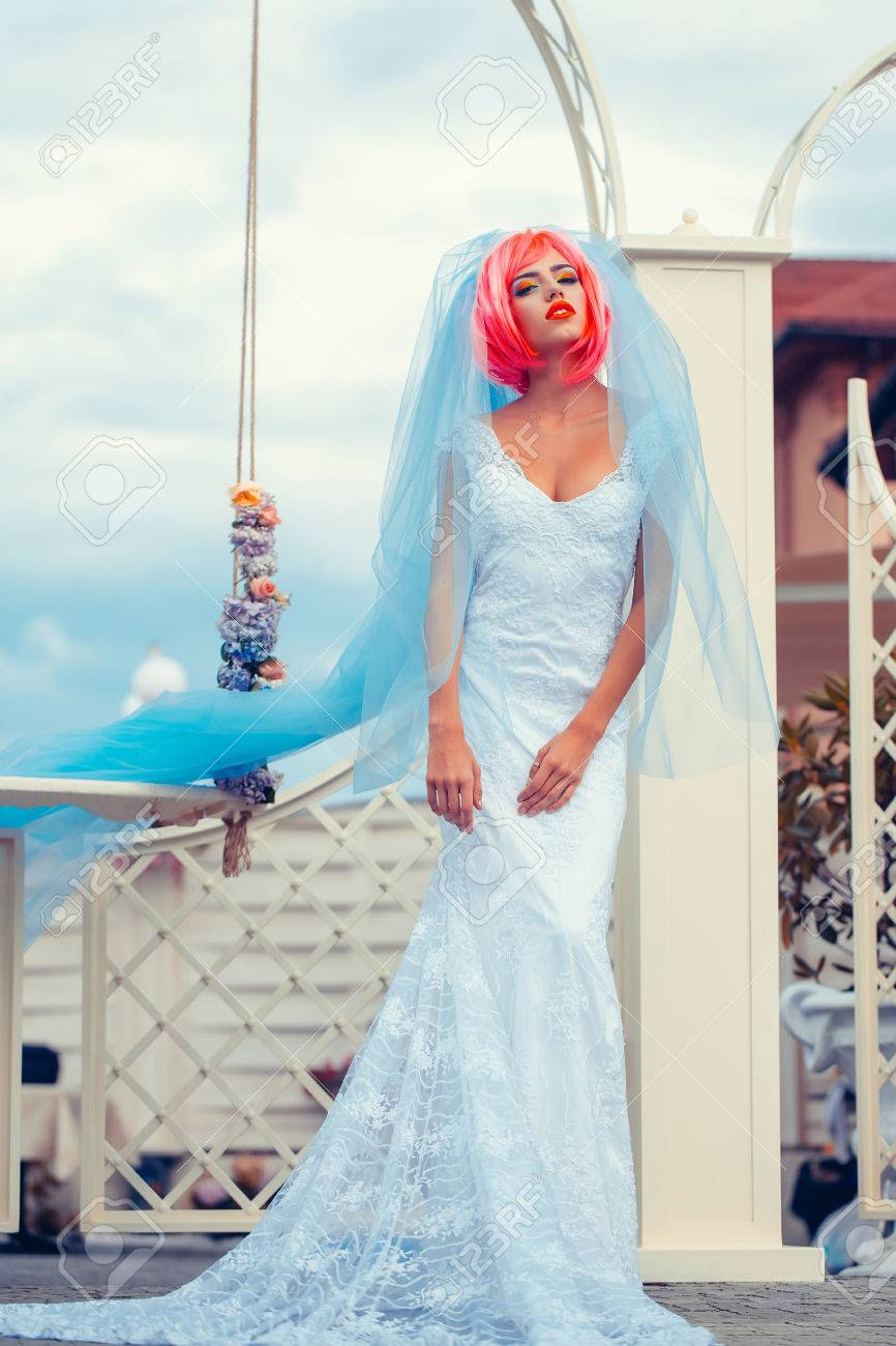 オレンジやピンクの髪と白いウェディング ドレスできれいな顔に明るいメイクや花のスイングに近い自然な曇り空を背景に青い花嫁のベールを持つ若い女性 の写真素材 画像素材 Image