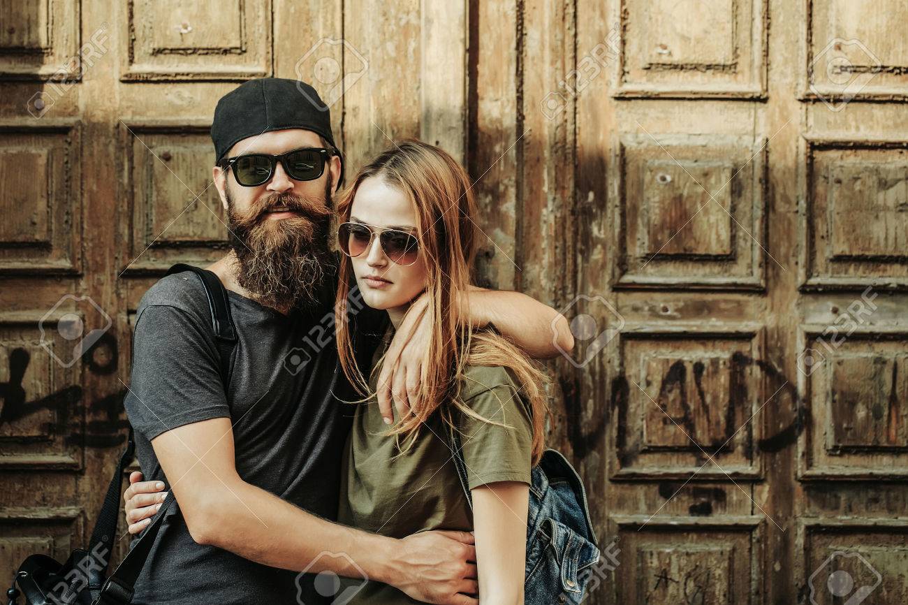 Young man hipster with beard on handsome face in sunglasses and baseball cap hugging pretty girl in casual clothes on wooden door background - 65086249 Young man hipster with beard on handsome face in sunglasses and baseball cap hugging pretty girl in casual clothes on wooden door background - 65086249