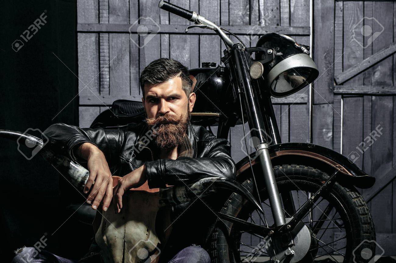 Bearded man hipster biker brutal male with beard and moustache in leather jacket sits on floor near motorcycle with bone skull antlers on wooden background - 67317755 Bearded man hipster biker brutal male with beard and moustache in leather jacket sits on floor near motorcycle with bone skull antlers on wooden background - 67317755