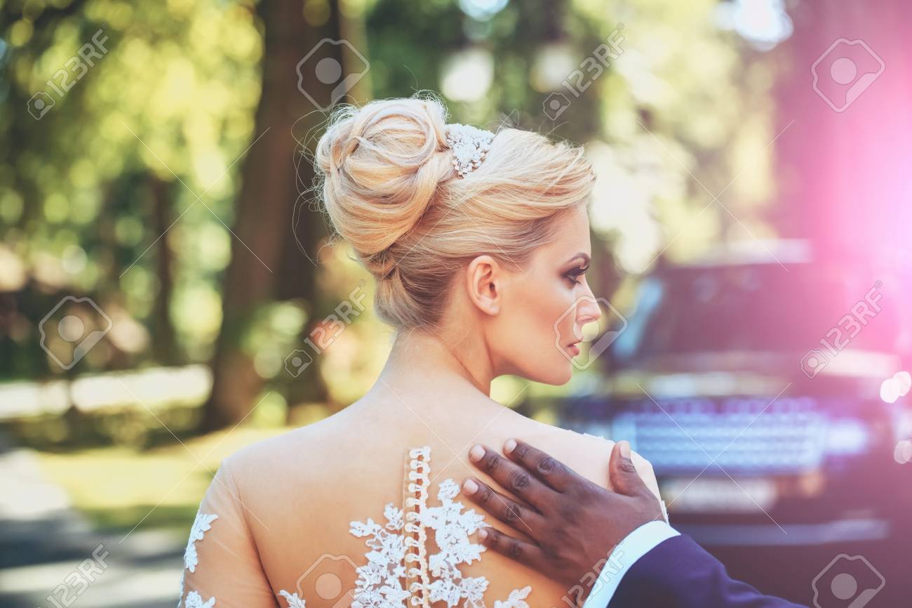 きれいな女の子やキレイなメイクとブロンドの髪 ヘアスタイルに自然な背景の日当たりの良い夏の日に白いセクシーなウェディング ドレスの肩にアフリカ系 アメリカ人の男性の手でかわいい花嫁 の写真素材 画像素材 Image