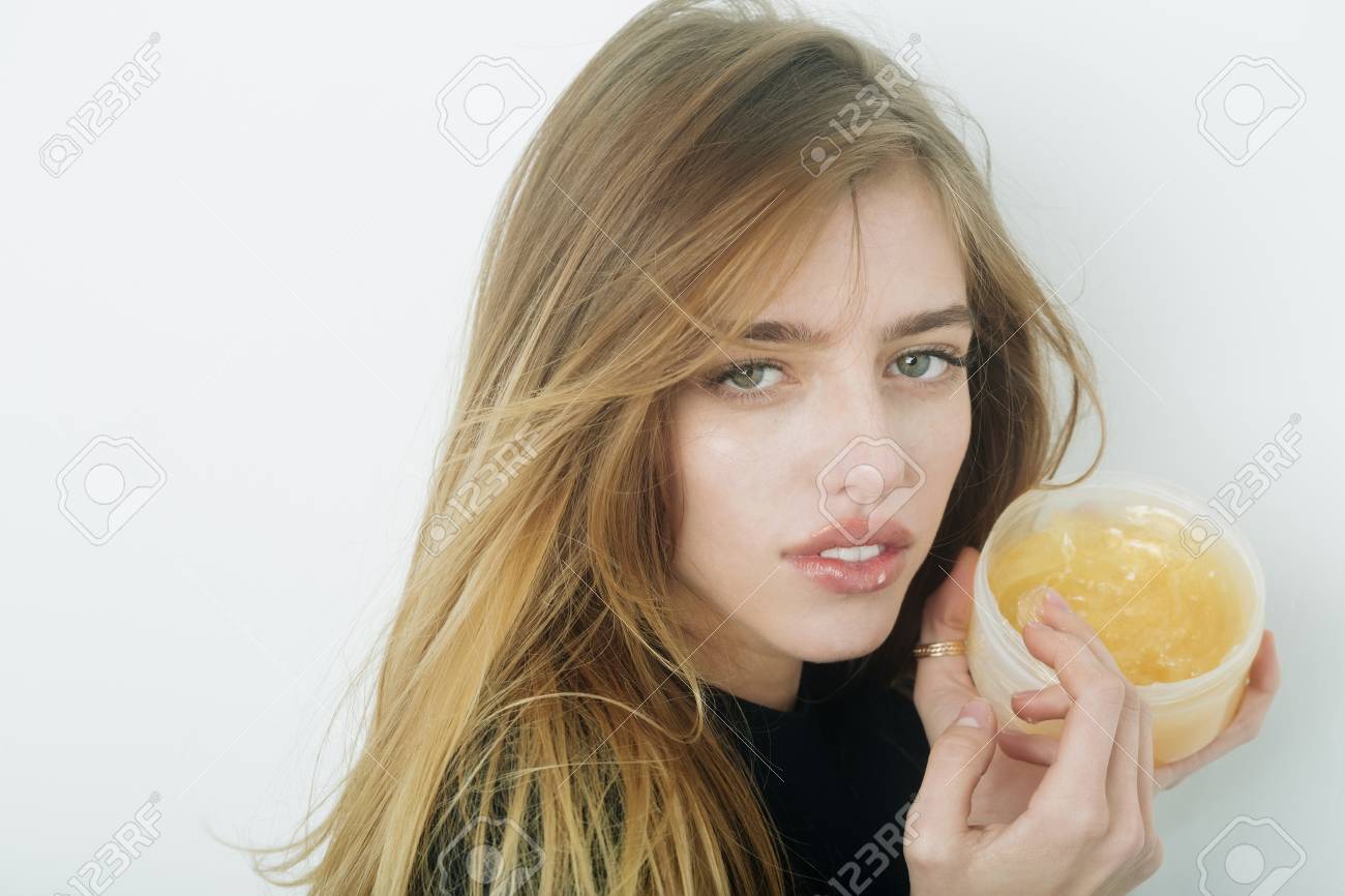 Hübsches Mädchen oder schöne Frau mit jungen Gesicht Haut und lange, blonden Haaren, ein Gel oder Balsam auf sexye Lippen aus Glas auf weiße Wand Hübsches Mädchen oder schöne Frau mit jungen Gesicht Haut und lange, blonden Haaren, ein Gel oder Balsam auf sexye Lippen aus Glas auf weiße Wand