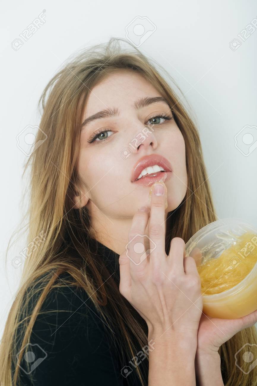 Hübsches Mädchen oder schöne Frau mit jungen Gesicht Haut und lange, blonden Haaren, ein Gel oder Balsam auf sexye Lippen aus Glas auf weiße Wand Hübsches Mädchen oder schöne Frau mit jungen Gesicht Haut und lange, blonden Haaren, ein Gel oder Balsam auf sexye Lippen aus Glas auf weiße Wand