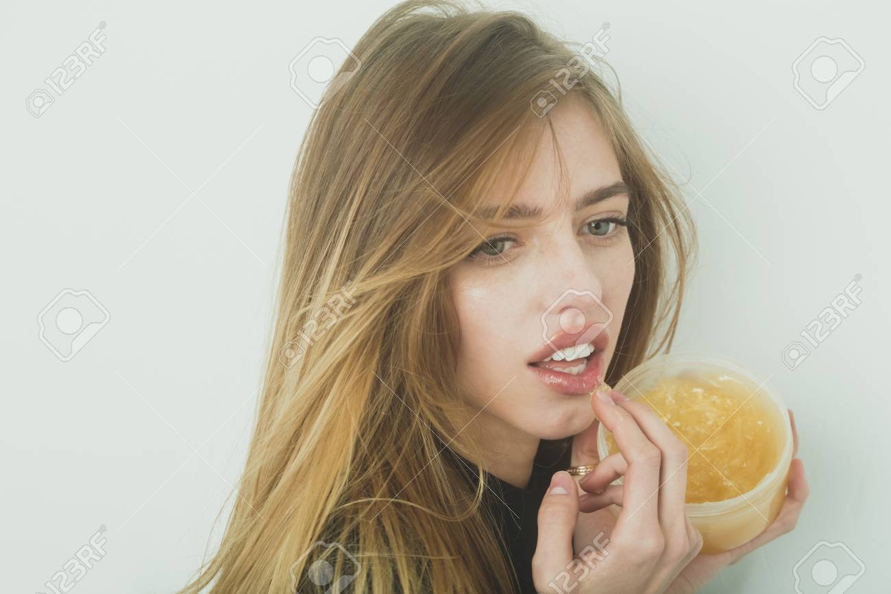 Hübsches Mädchen oder schöne Frau mit jungen Gesicht Haut und lange, blonden Haaren, ein Gel oder Balsam auf sexye Lippen aus Glas auf weiße Wand Hübsches Mädchen oder schöne Frau mit jungen Gesicht Haut und lange, blonden Haaren, ein Gel oder Balsam auf sexye Lippen aus Glas auf weiße Wand