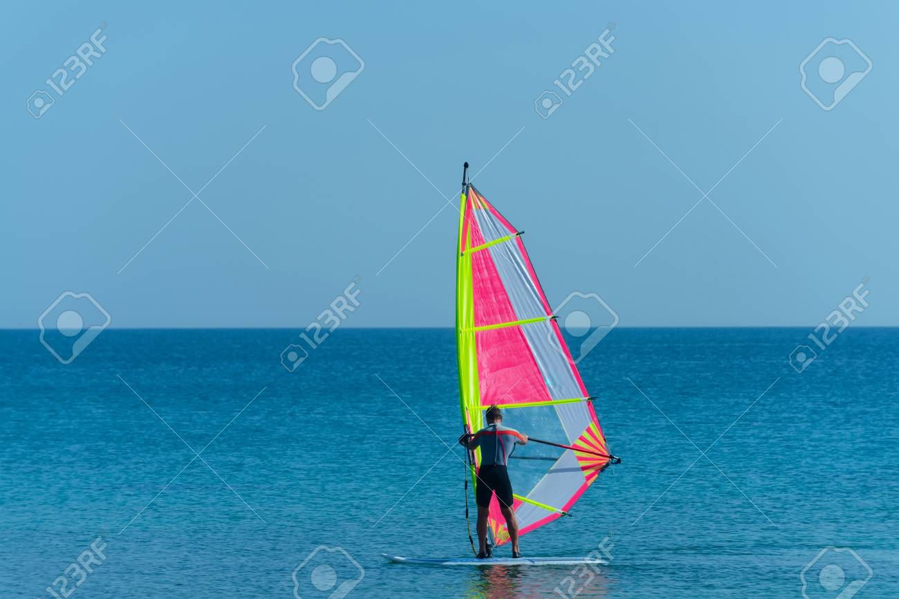 Windsurfer O Hombre Muscular El Traje De Windsurf A Con La Vela Colorida En El Mar O El Océano Tranquilo El Día En Del Cielo Azul. Deportes Acuáticos