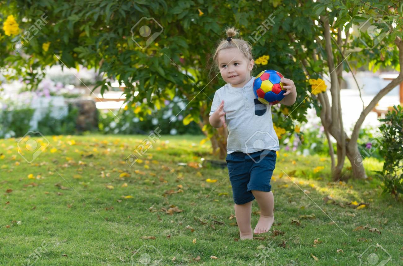 Sport And Vacation Cute Baby Boy Happy Little Child With Long Blond Hair Ponytail Barefoot Playing With Colorful Ball On Green Grass In Garden Sunny Summer Day On Natural Background Stock Photo