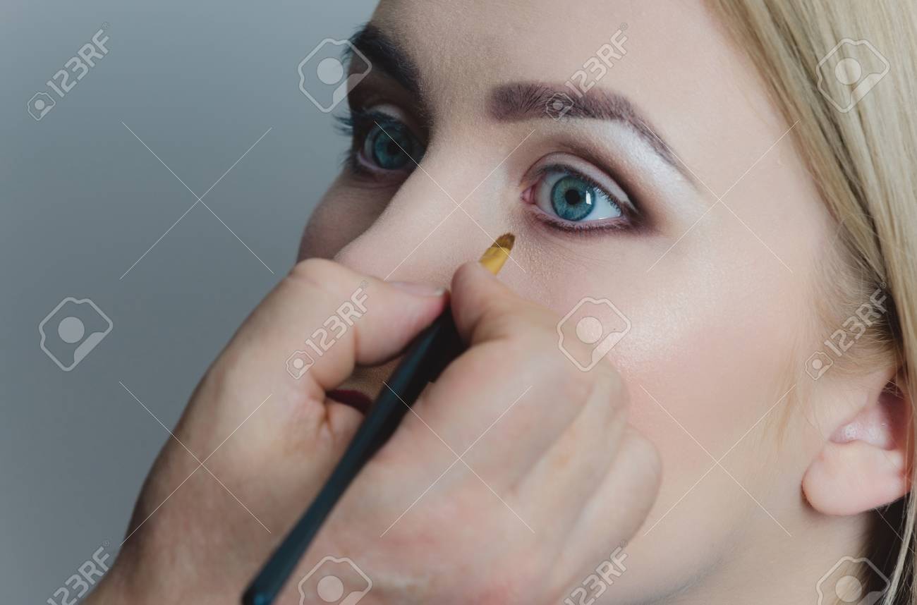 Femme Aux Yeux Bleus Se Maquiller Sur Fond Gris Main De Visagiste Mâle Appliquant Eyeliner Sur Le Visage De La Fille Avec Une Peau Saine Et Fond De