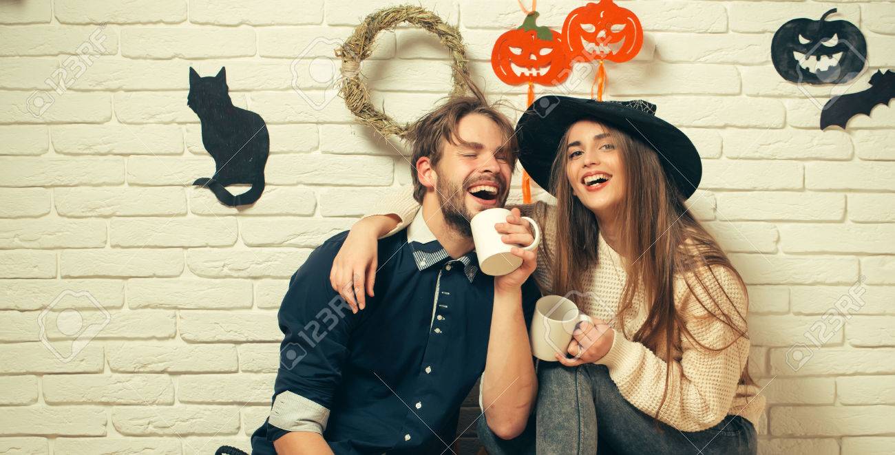 Halloween woman with long hair in witch hat. Happy couple in love with cups smiling. Man in casual wear sitting on floor. Holiday traditional symbols on white brick wall. Celebration and party concept - 84366260 Halloween woman with long hair in witch hat. Happy couple in love with cups smiling. Man in casual wear sitting on floor. Holiday traditional symbols on white brick wall. Celebration and party concept - 84366260