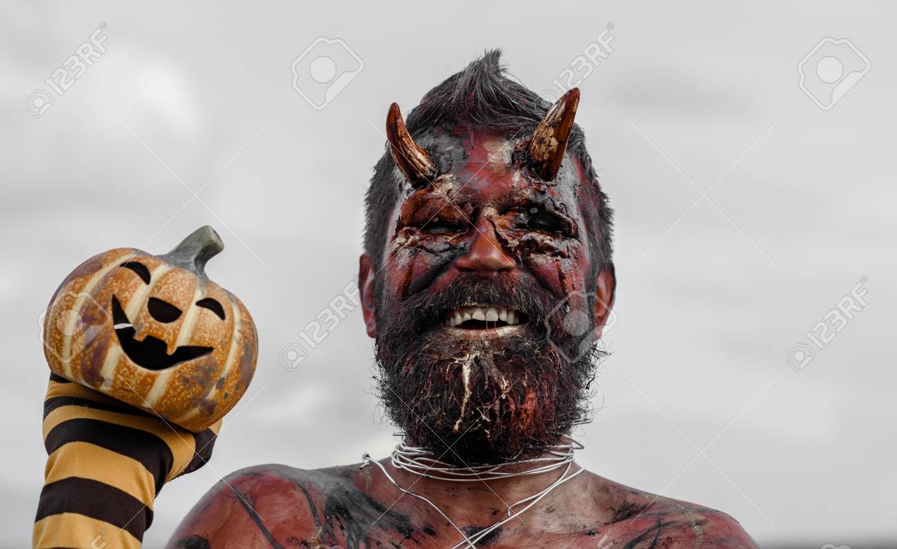 ハロウィーン悪魔ひげ笑みを浮かべて 赤い血の顔に傷します 頭に血の角を持つ悪魔 ストライプ靴下にカボチャを保持している魔人 悪 恐怖 地獄の概念 ジャック オ ランタンを持つドラゴン の写真素材 画像素材 Image