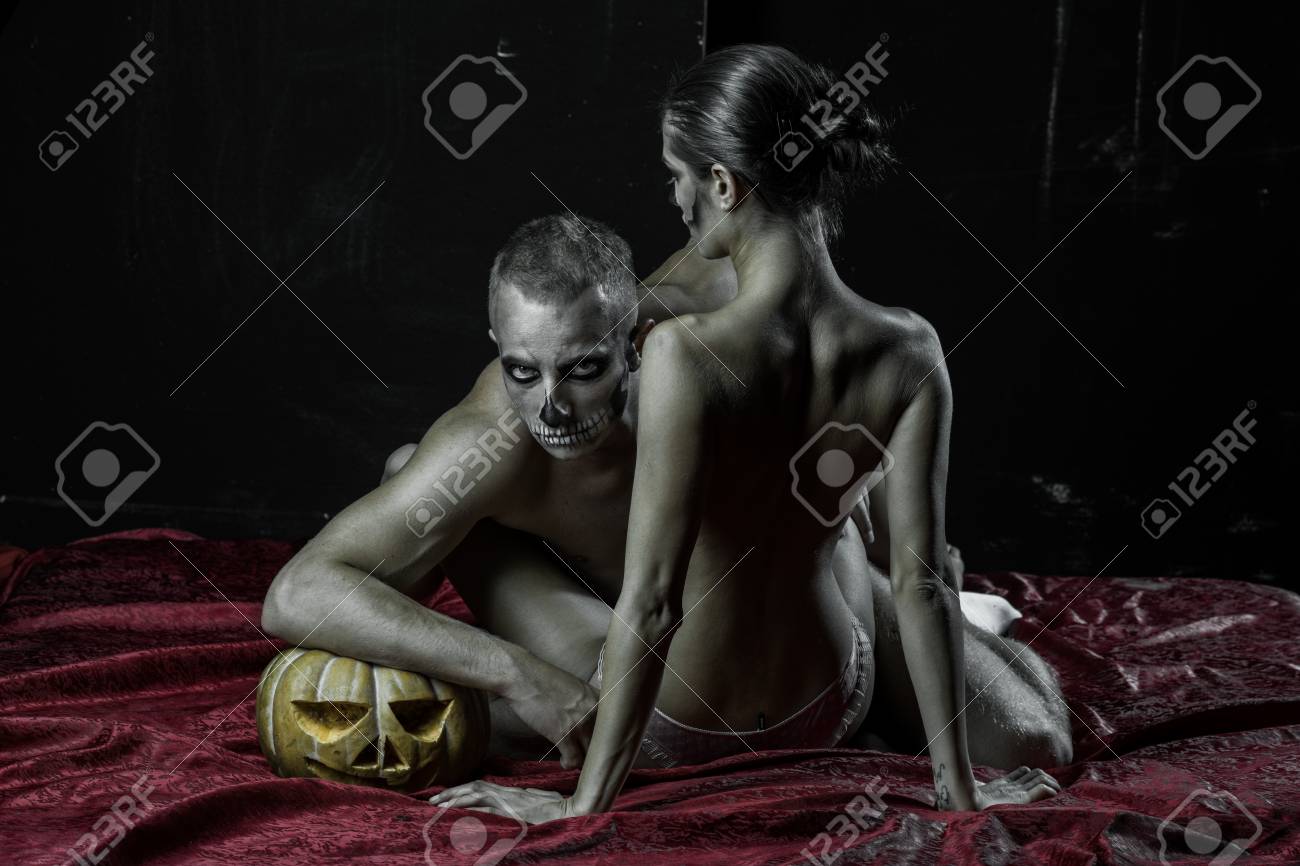 Halloween Holiday And Celebration. Halloween Couple With Makeup On Red  Bedcover. Man And Sexy Woman With Scary Face Embrace On Bed. Couple In Love  With Naked Body At Pumpkin. Zombie Party And