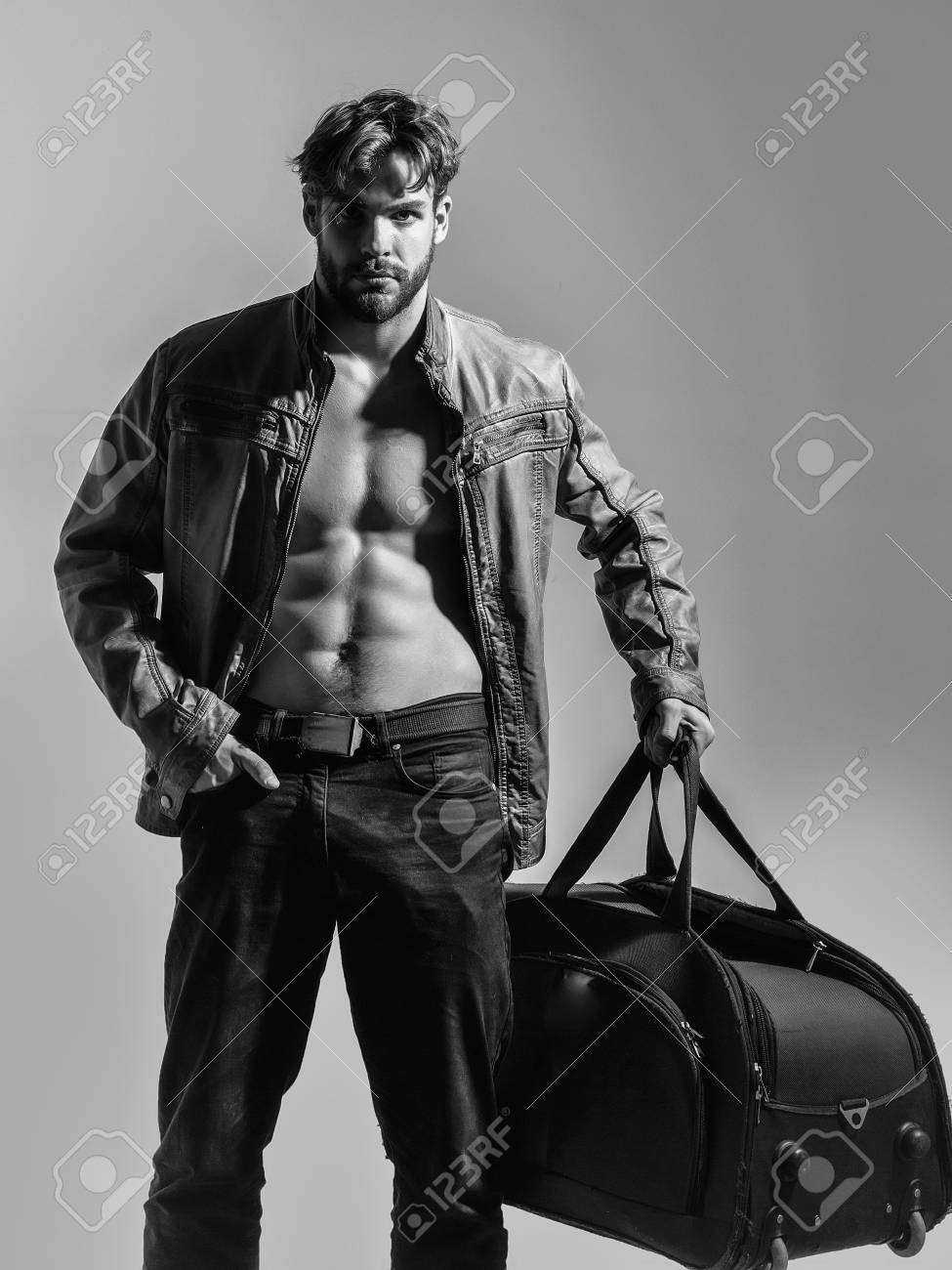 裸の胴体とセクシーなボディを持つイケメンの筋肉の男やひげの男らしく男は 灰色の背景にファッショナブルな茶色の革のジャケットとズボンに大きなバッグやスーツケースを保持しています の写真素材 画像素材 Image