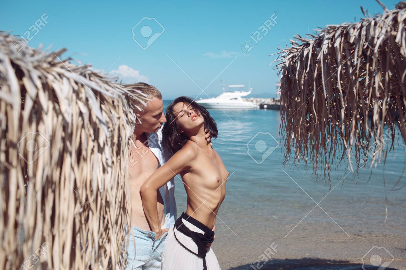 purenudism  beach Couple In Love Full Of Desire At Beach, Sea Background. Topless Woman Puts  Hand In Male Pants Near Umbrella Made Out Of Dried Palm Leaves. Couple On  Honeymoon At Tropical Seashore. Sex