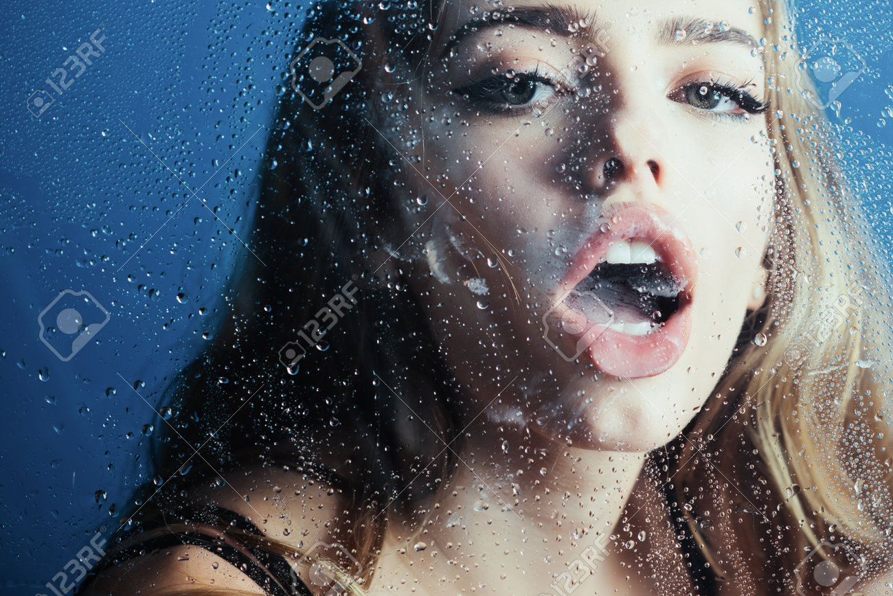 Sensual woman behind rain-covered window, water drops on glass, erotic dreamy expression in blue light