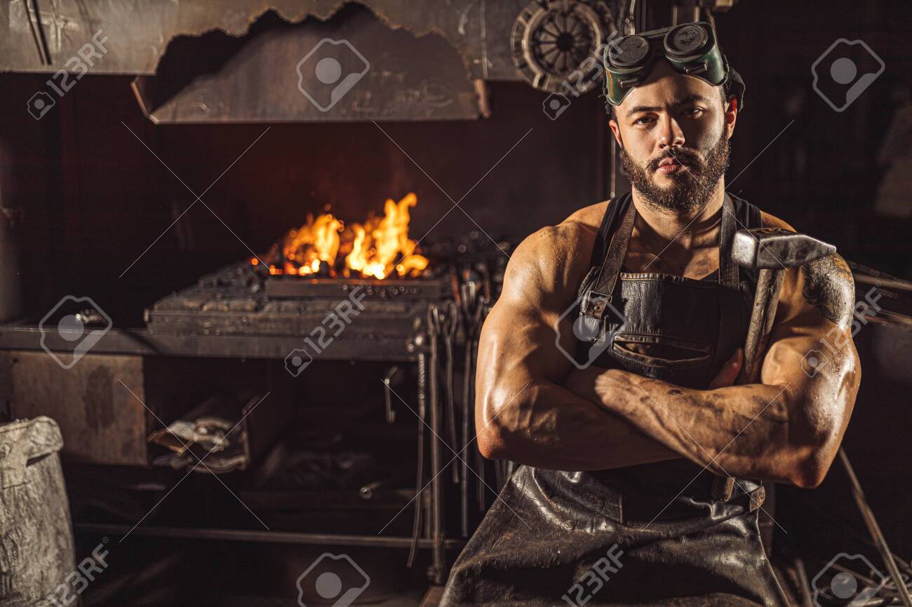Portrait of serious confident strong young metalworker in black apron stand looking at camera, work in workshop - 135675953 Portrait of serious confident strong young metalworker in black apron stand looking at camera, work in workshop - 135675953