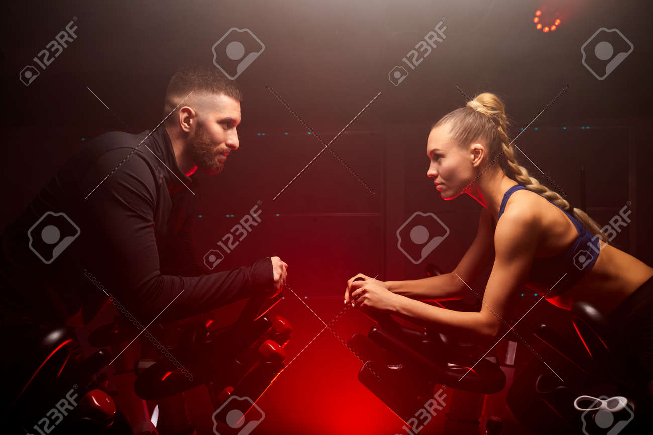 male and female sport people hold competition on bike in gym isolated on red neon lighted smoky space, oppsite of each other - 169212149 male and female sport people hold competition on bike in gym isolated on red neon lighted smoky space, oppsite of each other - 169212149