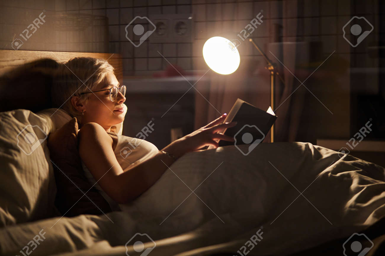 Beautiful female in pajamas and eyeglasses lying on bed reading book, alone at night. Caucasian short haired lady in bedroom, charming cute woman in room lighted by lamp - 169368811 Beautiful female in pajamas and eyeglasses lying on bed reading book, alone at night. Caucasian short haired lady in bedroom, charming cute woman in room lighted by lamp - 169368811
