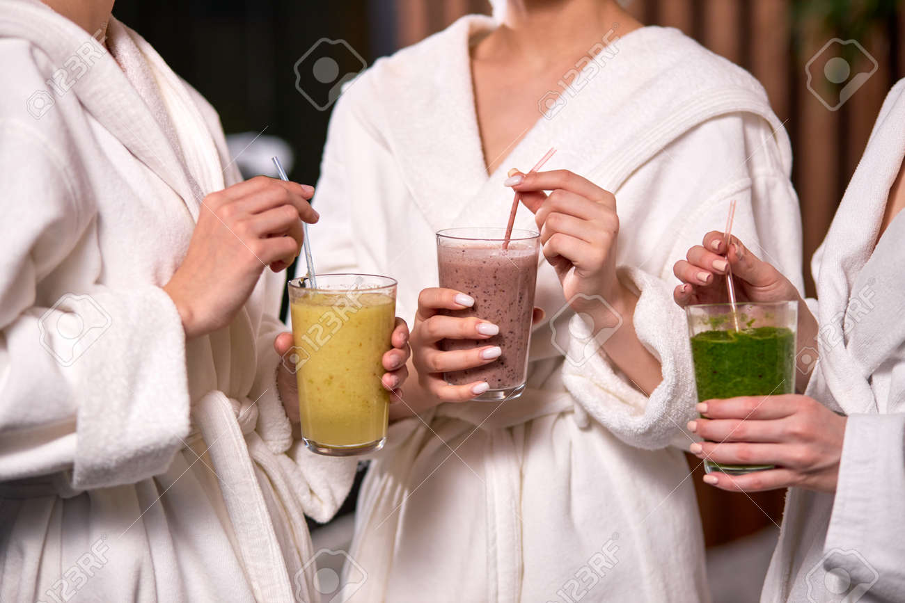 Cropped three women enjoy drinking healthy beverage during spa treatment wearing bathrobes, drinking freshly squeezed juice while spending weekend in wellness center. - 169369050 Cropped three women enjoy drinking healthy beverage during spa treatment wearing bathrobes, drinking freshly squeezed juice while spending weekend in wellness center. - 169369050