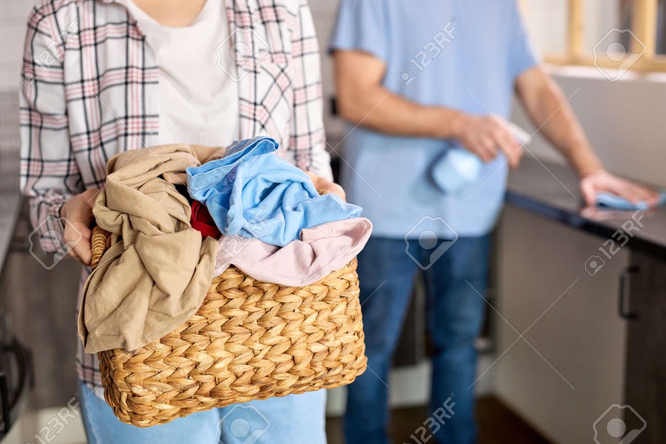 Casually dressed woman in checkered shirt holding basket full of dirty laundry needing washing, man cleaning kitchen in the background. Couple at home doing household chores, housekeeping - 169369338 Casually dressed woman in checkered shirt holding basket full of dirty laundry needing washing, man cleaning kitchen in the background. Couple at home doing household chores, housekeeping - 169369338