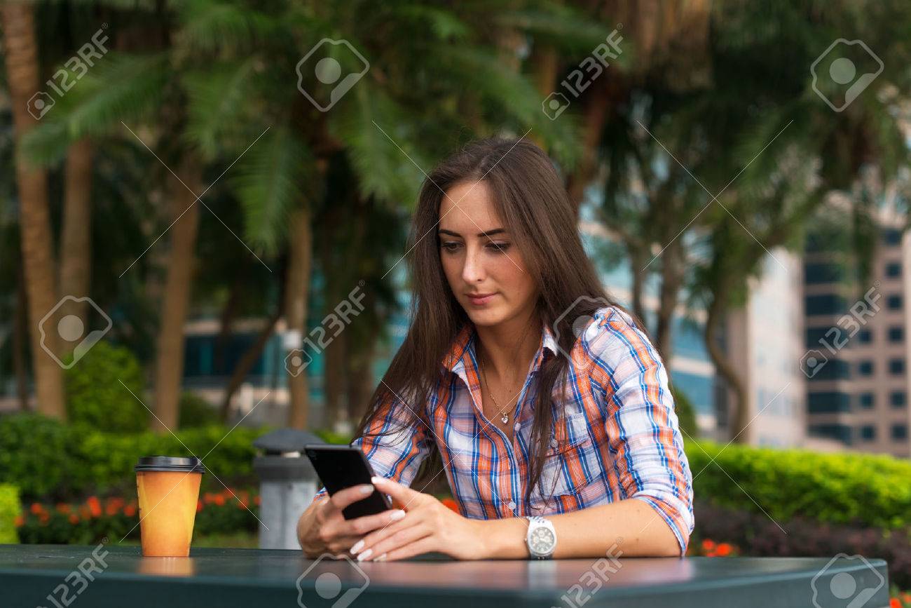 Young woman sitting outdoors reading and typing messages on her smartphone. - 80615726 Young woman sitting outdoors reading and typing messages on her smartphone. - 80615726