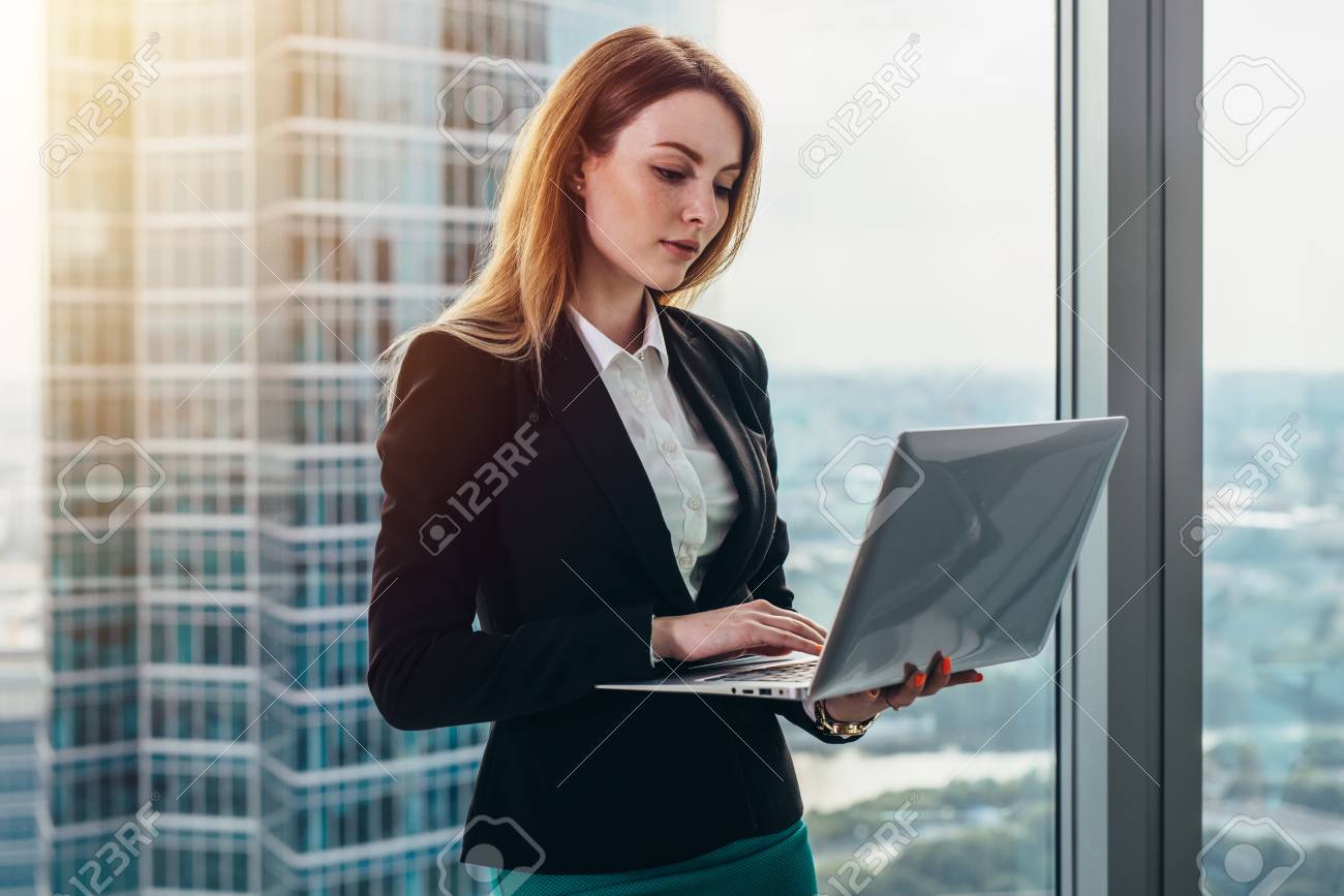 Young female lawyer working in her luxurious office holding a laptop standing against panoramic window with a view on business district - 89095730 Young female lawyer working in her luxurious office holding a laptop standing against panoramic window with a view on business district - 89095730