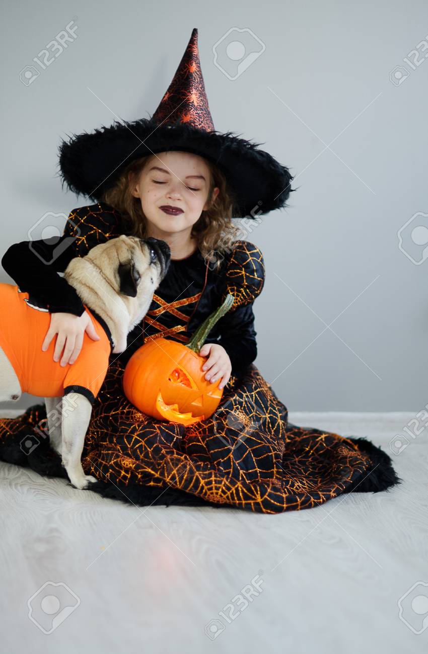 Petite Fille Et Son Amie - Carlin Sont Habillés En Costumes D'halloween.  Fille Dans Une Image De Fée Maléfique Est Assis Sur Un Sol. Sur Un Toutou A  Mis Un Pull Orange.