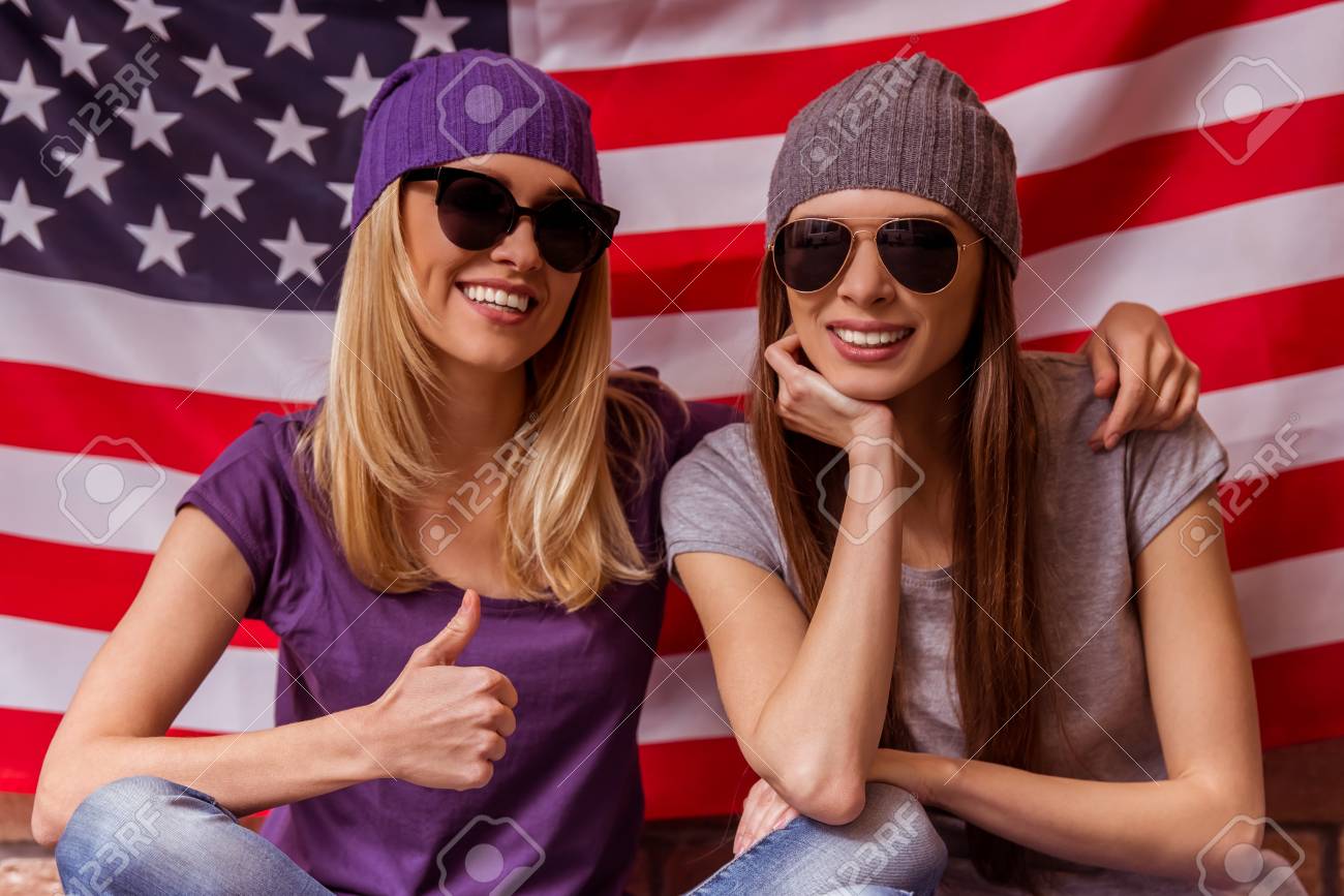 カジュアルな服とサングラス ポーズ 笑顔と Ok アメリカ国旗に対してあぐらを示す 2 つの美しい若い女の子 の写真素材 画像素材 Image