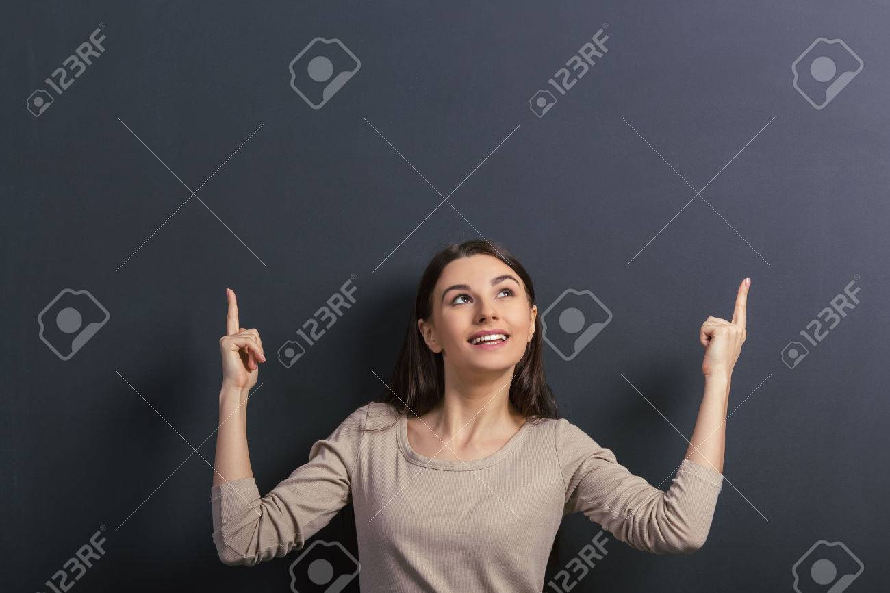 Beautiful young female student is smiling, looking and pointing up, standing against blackboard - 54005885 Beautiful young female student is smiling, looking and pointing up, standing against blackboard - 54005885