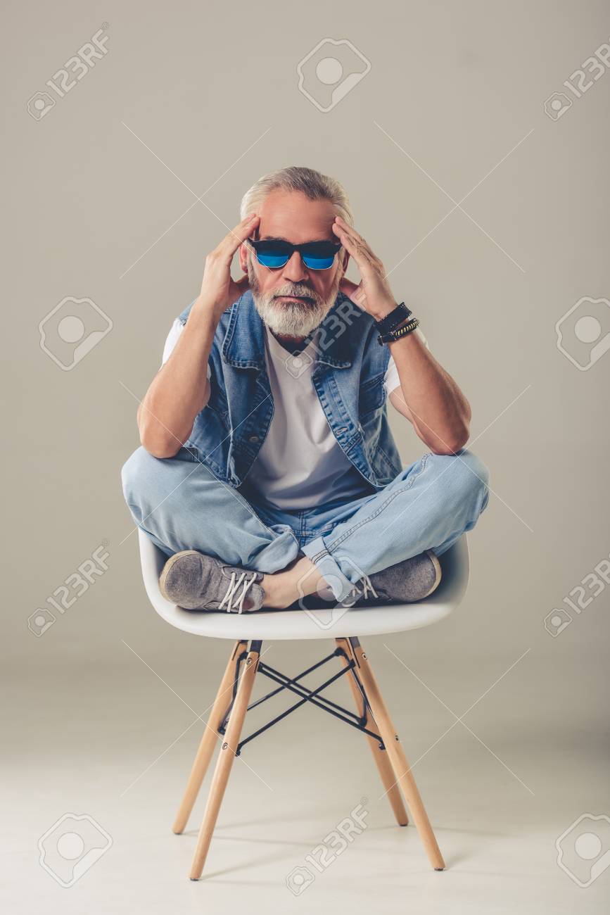 Hombre Maduro Con Barba Con Estilo En Jeans Y Gafas De Sol Está Mirando La Cámara Mientras Está Sentado En Una Silla Sobre Fondo Retratos, Imágenes Y Fotografía De