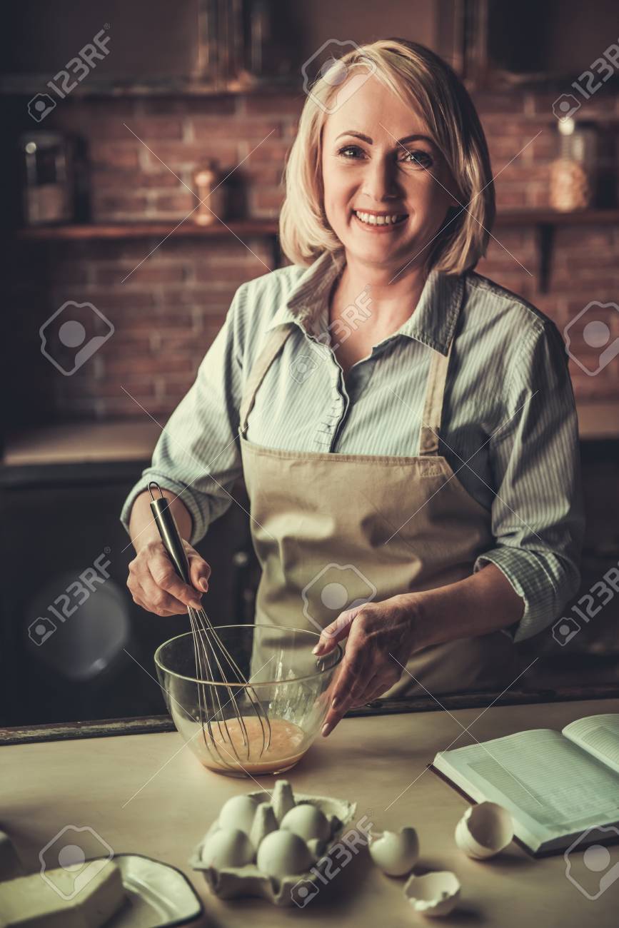 エプロンの中 美しい熟女の卵を Whisking カメラ目線 キッチンで焼きながら笑みを浮かべて の写真素材 画像素材 Image