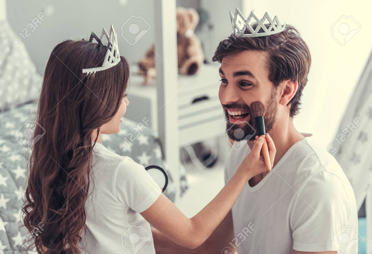 かわいい小さな娘と王冠で彼女のハンサムな若いお父さんは子供の部屋で一緒に遊んでいます 女の子は化粧彼女のお父さんをやっています の写真素材 画像素材 Image