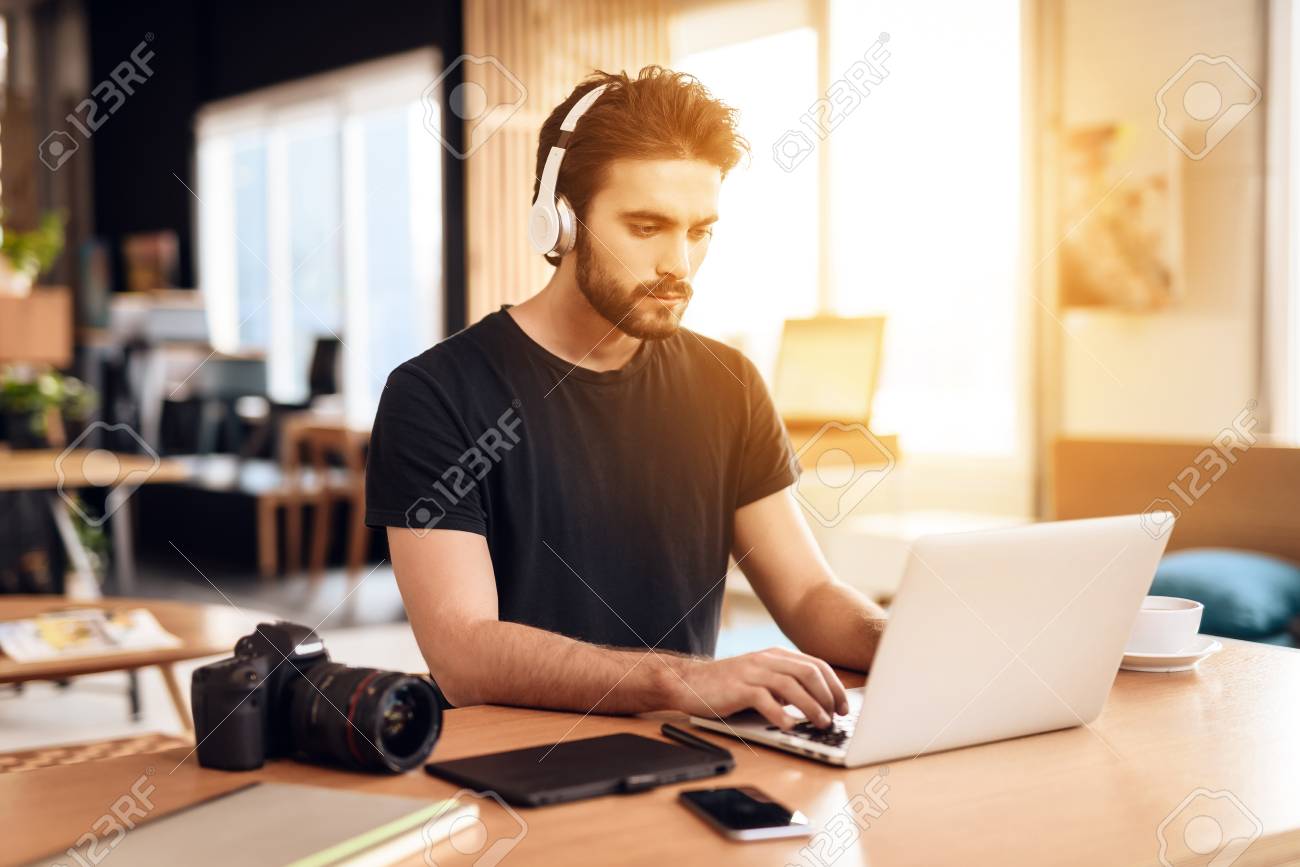 机に座っているノートパソコンで音楽を聴くtシャツを着たフリーランサーのひげを生やした男 の写真素材 画像素材 Image