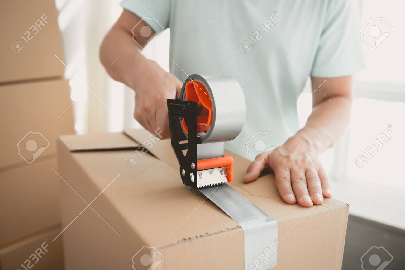 Close up. Packing Boxes with Building Tape in Order to Move to New Housing. Moving the Family to New Place. Family Gathers Things in Cardboard Boxes. Young Man in Room. New Apartment Concepts. - 105986034 Close up. Packing Boxes with Building Tape in Order to Move to New Housing. Moving the Family to New Place. Family Gathers Things in Cardboard Boxes. Young Man in Room. New Apartment Concepts. - 105986034