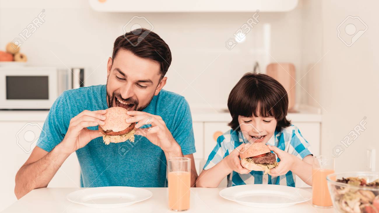 Smiling Son and Father Have Lunch in Kitchen. Boy in Shirt. Modern Kitchen. Sitting Boy. Boy with Spoon. Breakfast in Morning. White Table in Kitchen. Burger in Hands. Young Father. - 115669036 Smiling Son and Father Have Lunch in Kitchen. Boy in Shirt. Modern Kitchen. Sitting Boy. Boy with Spoon. Breakfast in Morning. White Table in Kitchen. Burger in Hands. Young Father. - 115669036