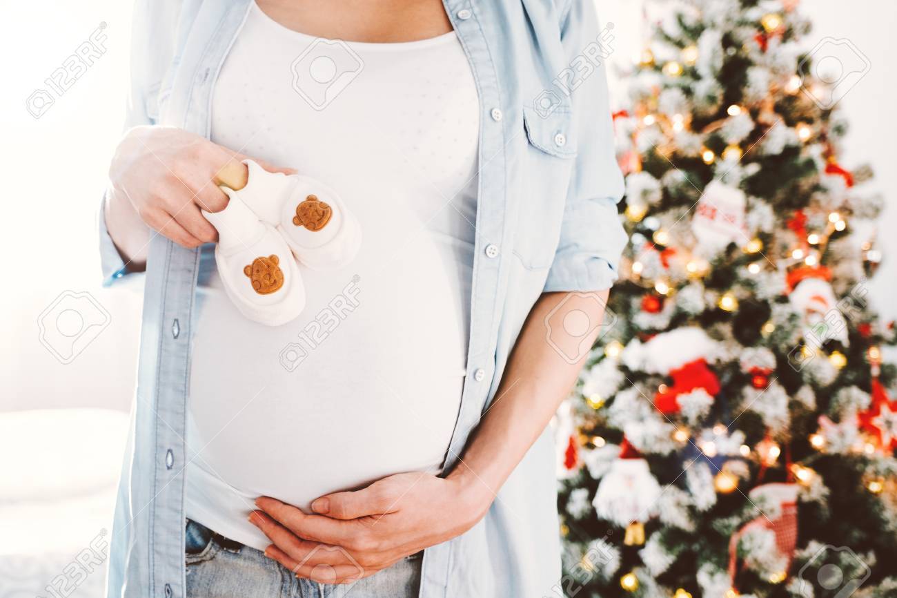 Happy beautiful pregnant woman holds Cute little infants shoes in hands at christmas tree in bright white room and touching her belly. - 87678200 Happy beautiful pregnant woman holds Cute little infants shoes in hands at christmas tree in bright white room and touching her belly. - 87678200