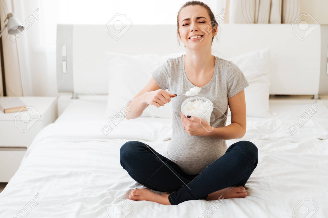 座っていると寝室でベッドの上にアイスクリームを食べる動揺妊娠中の若い女性 の写真素材 画像素材 Image