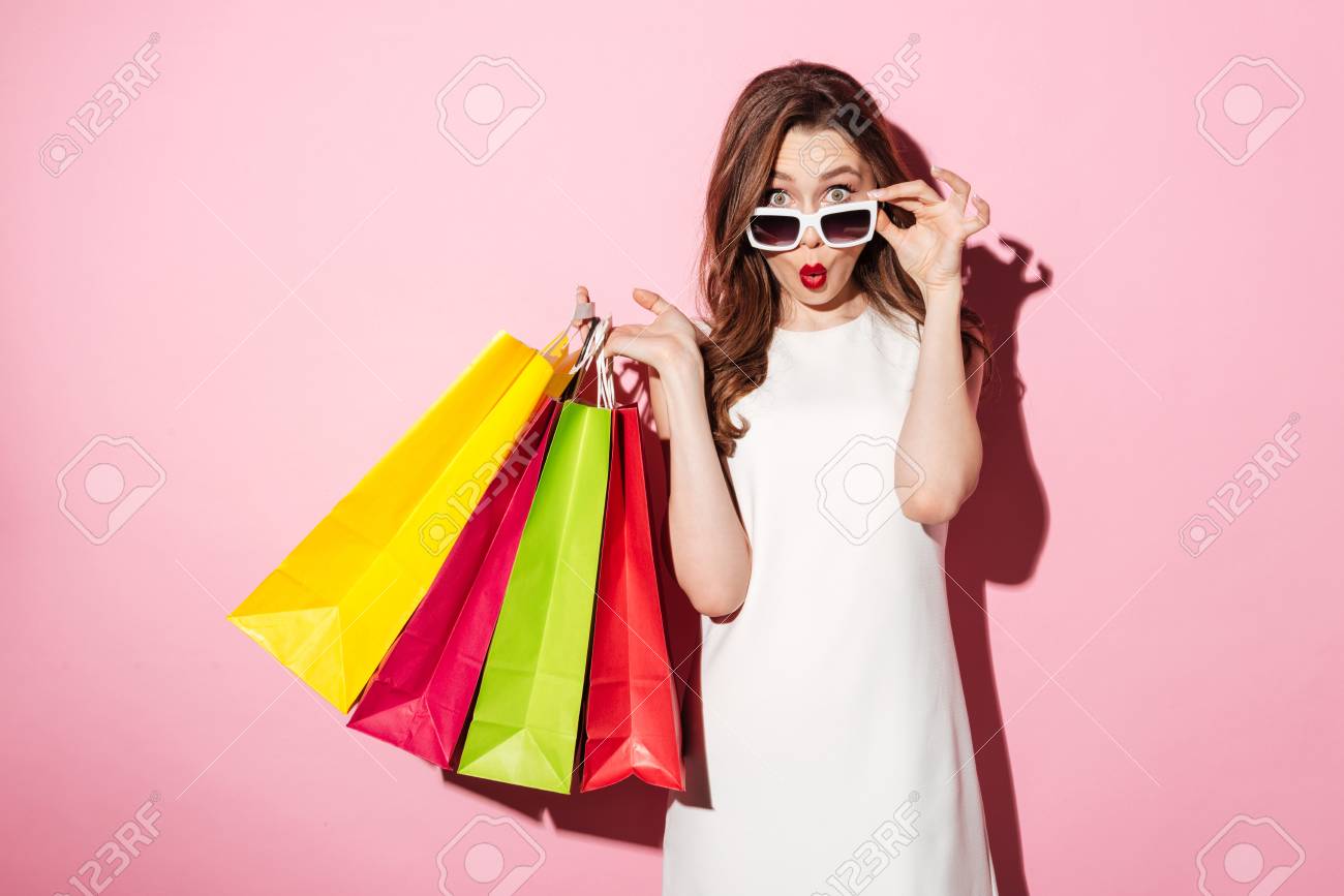 Image of a shocked young brunette lady in white summer dress wearing sunglasses posing with shopping bags and looking at camera over pink background. - 80853874 Image of a shocked young brunette lady in white summer dress wearing sunglasses posing with shopping bags and looking at camera over pink background. - 80853874