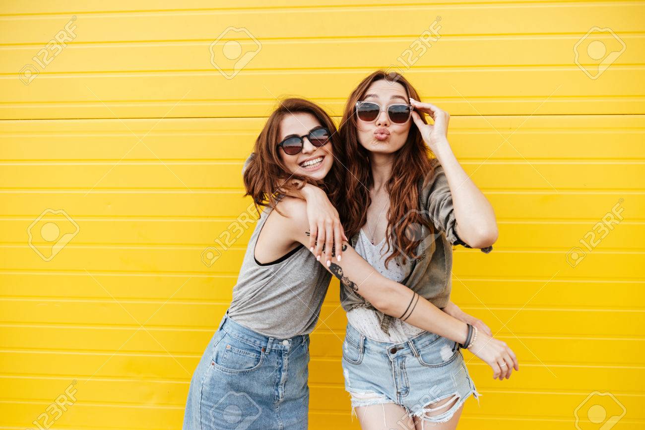 Image of two young happy women friends standing over yellow wall. Looking at camera blowing kisses. - 84291185 Image of two young happy women friends standing over yellow wall. Looking at camera blowing kisses. - 84291185